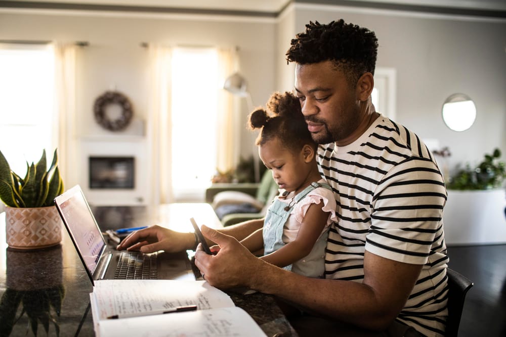 New dad working from home while holding his daughter in his lap, thinking, "Preparing for fatherhood is impossible."